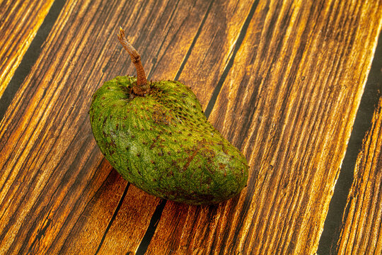 Green soursop fruit with spiky skin and brown stem resting on rustic wooden surface with warm golden tones