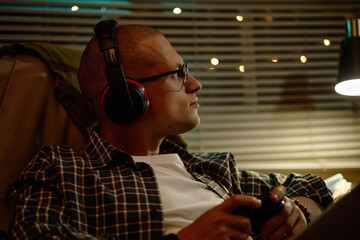Young adult Caucasian man wearing headphones and holding gamepad while playing video games at night. Visual metaphor for esports, digital entertainment, gaming addiction campaigns © Seventyfour