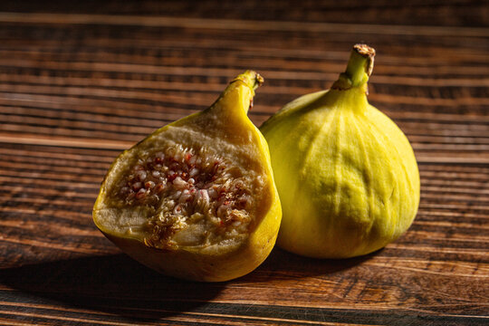 Fresh yellow fig cut in half next to whole fig on dark rustic wood surface with dramatic side lighting and shallow depth of field