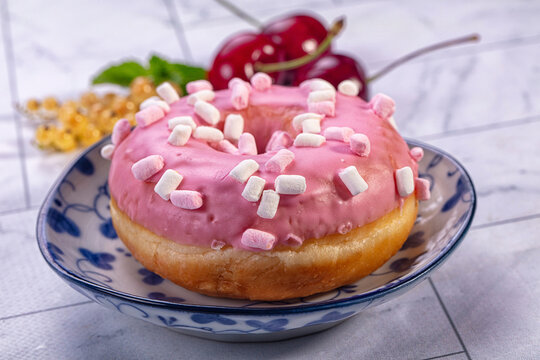 Pink glazed donut topped with mini marshmallows on blue floral plate, fresh cherries and mint in soft focus background