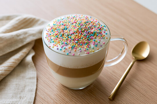 Glass cup filled with layered coffee topped with whipped cream and colorful sprinkles, placed on a wooden table beside a golden spoon and a beige cloth