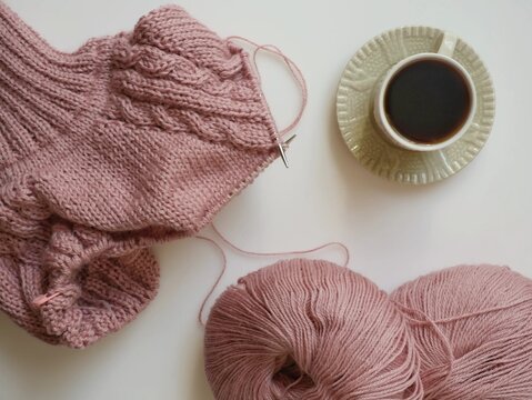 Top view flat lay of unfinished pink knitted sweater on circular needles, skeins of yarn and cup of coffee on white background, cozy knitting hobby and autumn craft concept