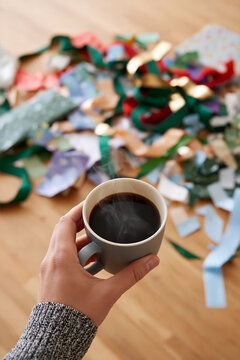 Hand holding gray coffee cup with steaming black coffee above colorful wrapping paper and ribbons scattered on wooden floor in a cozy indoor setting