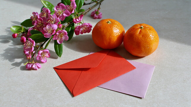 Two oranges placed beside red and pink envelopes with a bouquet of pink flowers on a light-colored surface, creating a vibrant and colorful arrangement