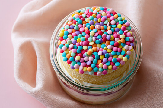 Colorful dessert with a layer of cream and a topping of vibrant sprinkles in a glass dish placed on a soft pink fabric background