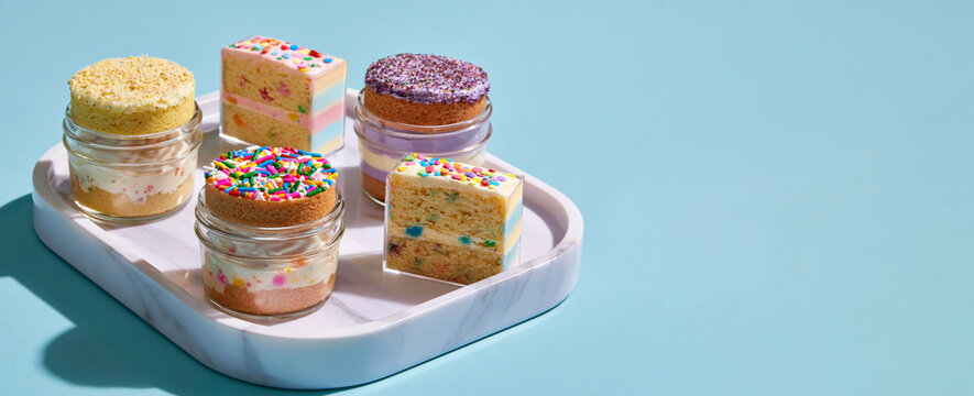 Colorful layered desserts in glass jars with sprinkles displayed on a marble tray against a light blue background, showcasing a variety of sweet treats