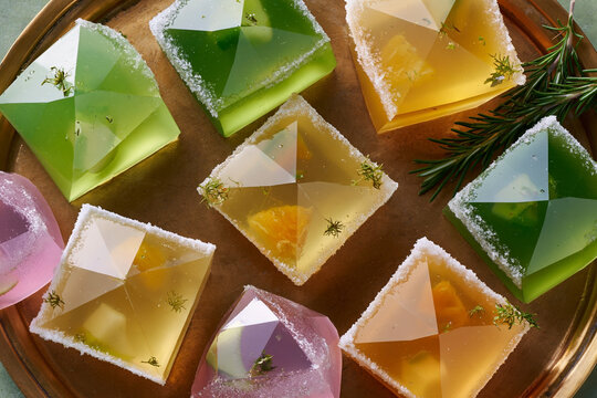 Colorful geometric gelatin desserts arranged on a golden tray with a sprig of rosemary, featuring various shapes and vibrant hues of green, yellow, and pink