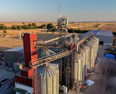 Aerial view of a grain silo facility with metal storage bins, a red industrial tower, and conveyor systems surrounded by agricultural fields during golden hour in Pancevo, Serbia.