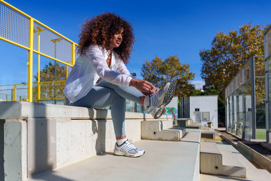Young woman with curly hair tying her shoelace on concrete steps, preparing for sports activity outdoors