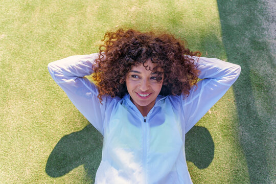 Woman with curly hair resting on artificial grass after exercise, feeling relaxed and happy, fitness lifestyle