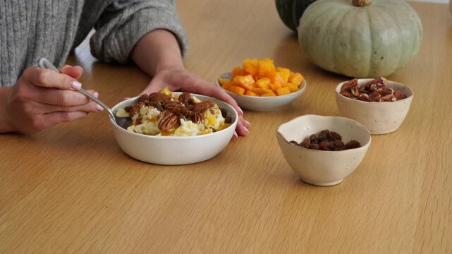 Hands scoop warm rice millet pudding porridge topped with pumpkin and pecans in cozy autumn kitchen, spoons and bowls on light wooden table with diced fruit, raisins and pumpkin, closeup