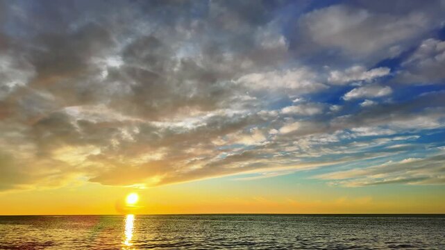 A golden sun sets over calm sea water beneath a dramatic layered cloudscape. Timelapse.