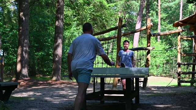 A candid moment of couple enjoying a friendly, amateur table tennis (ping-pong) match outdoors on a bright, sunny day with high-contrast lighting in a scenic forest setting.