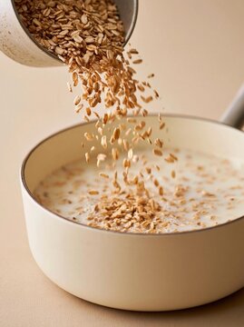 White bowl with a spoon in it. the bowl is filled with a creamy white substance, which appears to be oatmeal or porridge. the oatmeal is being poured from a white measuring cup into the bowl.