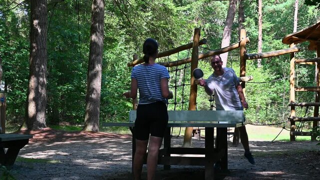 A candid moment of couple enjoying a friendly, amateur table tennis (ping-pong) match outdoors on a bright, sunny day with high-contrast lighting in a scenic forest setting.