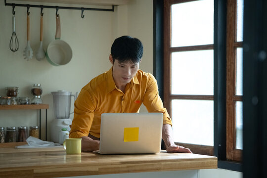 Young Asian Man Reading Morning News on Laptop in Kitchen with Coffee Mug