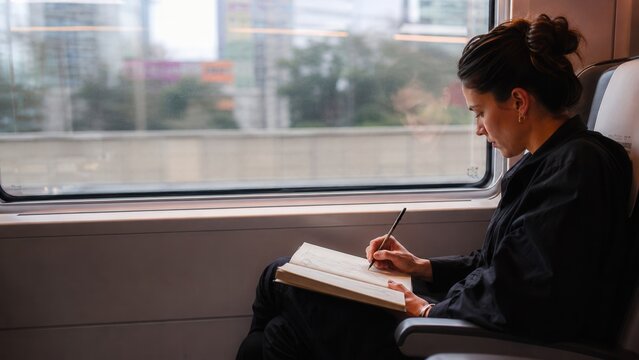 Woman drawing in sketchbook while traveling by train with city view through the window