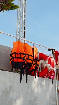 Vertical. Orange Life Jackets and a Red Lifebuoy Hang on a Rack Above a Concrete Wall Under Blue Sky, Supporting Water Safety and Rescue Preparedness