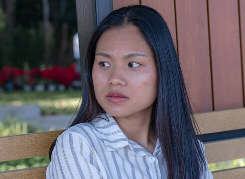 A thoughtful young Asian woman sitting on a wooden park bench outdoors, wearing a stylish striped shirt and looking away pensively with a calm expression against a beautiful background of green trees.