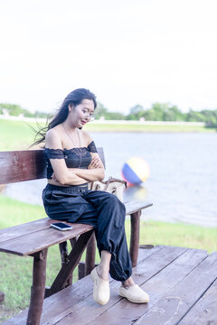 Young woman enjoying a peaceful moment by the lake, seated on a rustic wooden bench in casual fashion. Soft summer light, serene water, and lush greenery create a tranquil outdoor lifestyle scene.