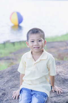 A cute young Asian boy sitting on a large rock by a calm lake with hands resting behind, wearing a yellow polo shirt and cute denim shorts, smiling happily outdoors on a sunny day with a beach ball.