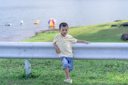 A cute young Asian boy wearing a yellow polo shirt and casual denim shorts leaning on a white metal guardrail by a serene reservoir, looking away on a bright sunny day with a colorful floating ball.