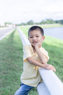 A charming portrait of a cute young Asian boy leaning on a white metal guardrail in a scenic park, capturing a sweet family lifestyle moment with warm natural lighting and a calm, gentle expression.