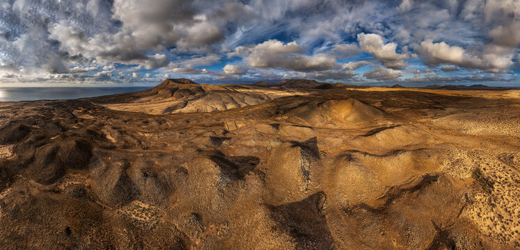 Aerial view of Parque Natural de Corralejo featuring volcanic terrain, rolling sand dunes, and distant mountains under a dramatic cloudy sky in Canary Islands, Spain.
