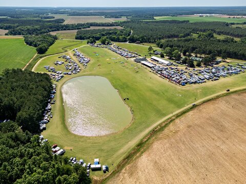 Aerial view of the Bad Apples ppg flyin event at Hodges Field with paragliders in the sky, numerous parked vehicles, a pond, and surrounding forest in Andersonville, Georgia, United States.