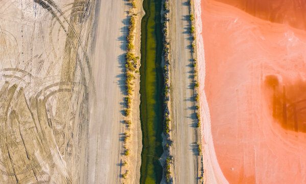 Aerial view of pink salt evaporation ponds and a green water channel, showing the geometric landscape of industrial sea salt production in the Camargue Aigues-Mortes, Occitanie, France.