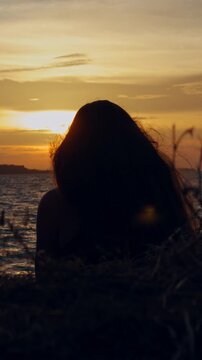 Vertical. Silhouetted Woman Sits Among Shoreline Grasses Facing Rippling Water and Golden Sunset, Evoking Nature and Quiet Lifestyle Vertical video.