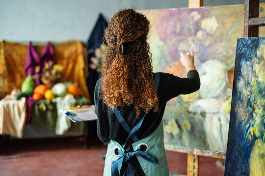 Teenage girl in college painting class, art studio, art workshop. She paints still life on canvas with oil paints, holds brush, palette.
