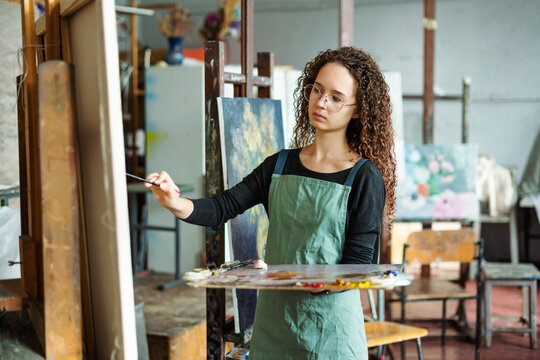 Portrait of teenage girl in painting class, art studio, art workshop. She paints still life on canvas with oil paints, holds brush, palette. Studying painting in college.