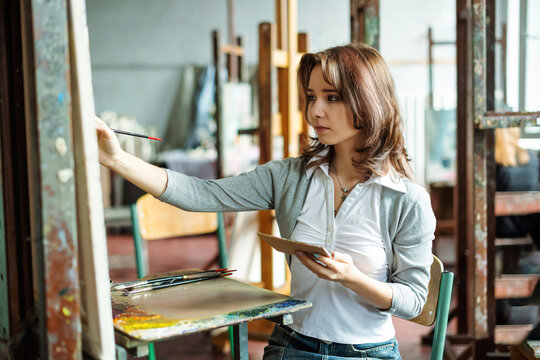 Portrait of teenage girl in painting class, art studio, art workshop. She paints still life on canvas with oil paints, holds brush, palette. Studying painting in college.