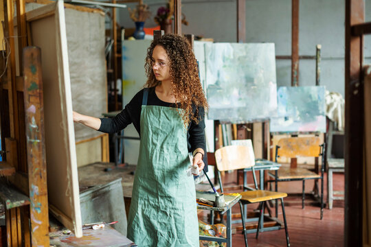 Portrait of teenage girl in painting class, art studio, art workshop. She paints still life on canvas with oil paints, holds brush, palette. Studying painting in college.