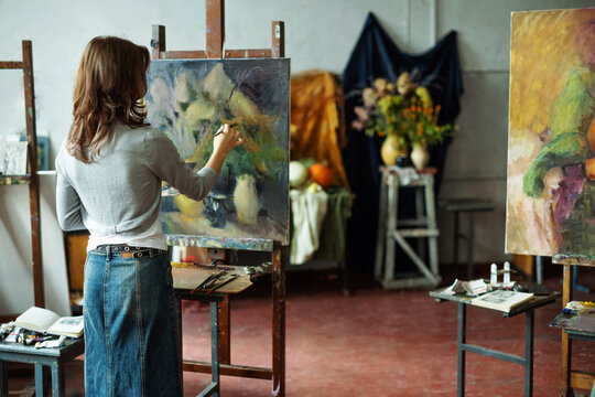 Teenage girl in painting class, art studio, art workshop. She paints still life on canvas with oil paints, holds brush, palette. Studying painting in college.