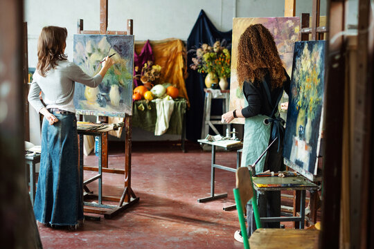 Two teenage girls in college painting class, painting still life on canvas with oil paints, holding brush, palette. Art studio, art workshop. Creativity development