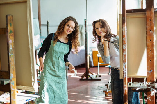 Two teenage girls in college painting class, painting still life on canvas with oil paints, holding brush, palette. Art studio, art workshop.