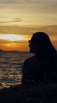 Vertical. Woman Silhouette Watches Golden Dusk over Ocean From Grassy Shore, Dark Sunglasses and Rippling Water Evoke Quiet Coastal Lifestyle Vertical video.