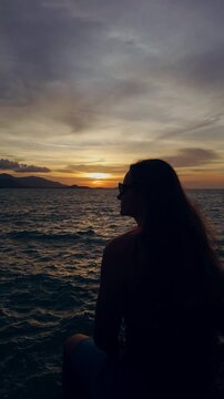 Vertical. Woman Wearing Sunglasses Sits Before an Ocean Sunset, Silhouetted Against Rolling Waves, Distant Islands and a Cloudy Coastal Sky Vertical video.