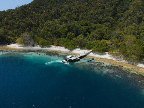 Aerial view of the Piaynemo landmark featuring a wooden pier with a boat docked in clear turquoise waters surrounded by lush tropical forest in Raja Ampat, Southwest Papua, Indonesia.