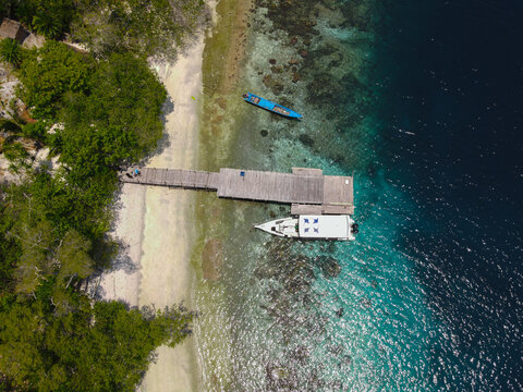 Aerial view of Piaynemo with a wooden pier, a white boat, and a blue canoe moored in the clear turquoise waters of a tropical beach in Raja Ampat, Southwest Papua, Indonesia.