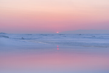 Obraz premium Pastel sunset over a frozen sea with snow-covered ice and distant hills, creating a calm and peaceful winter landscape.