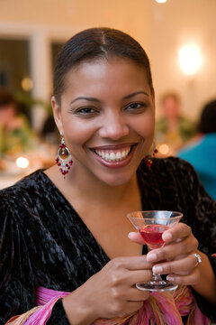 African American woman in elegant attire smiles while holding a cocktail glass at a festive gathering, with blurred guests and warm lighting in the background