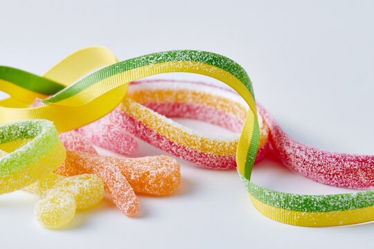 Colorful gummy candies in various shapes and sizes, including sour belts and jelly beans, arranged with vibrant ribbons on a white background