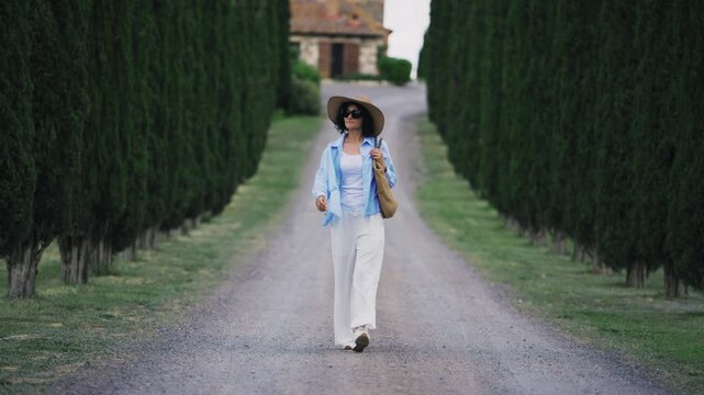 Stylish woman walking along cypress tree lined road in tranquil Tuscany countryside