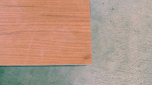 High-angle shot of a smooth wooden surface corner placed on a textured, rustic gray concrete floor, showing contrast between warm wood grain and cool industrial background with copy space.