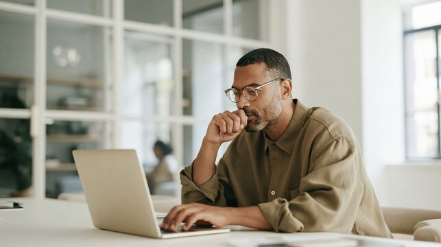 Professional man thinking at a laptop, a natural, analytical moment in a bright, glass-walled office with soft diffuse light for business and remote consulting campaigns, featuring copy space.