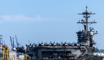Fighter jets line the aircraft carrier. Large cranes tower over the aircraft carrier. A military aircraft carrier rests at the harbor. © Nutchanart