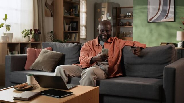 African American man shopping online using smartphone and credit card on sofa. Commercial clip suitable for mobile banking apps, fintech services and e-commerce campaigns